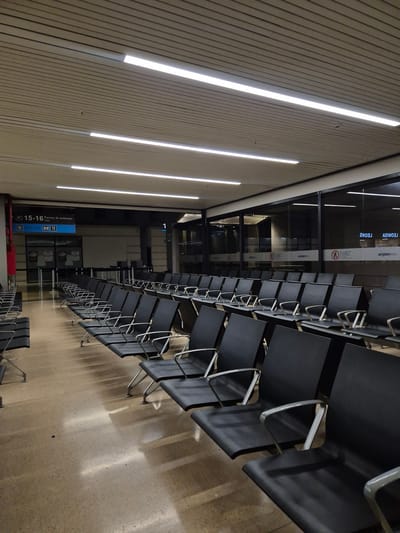 The image depicts a waiting area in an airport, specifically in Rionegro, Colombia, based on the signage visible in the background. Rows of empty black chairs are arranged along a wall with vertical light strips. The chairs are facing a glass wall with the word "airplan" visible on it. Signage for "gates" in Spanish is posted near the bottom of the image. The floor appears to be made of concrete or tile. The scene is deserted, with no people present.
