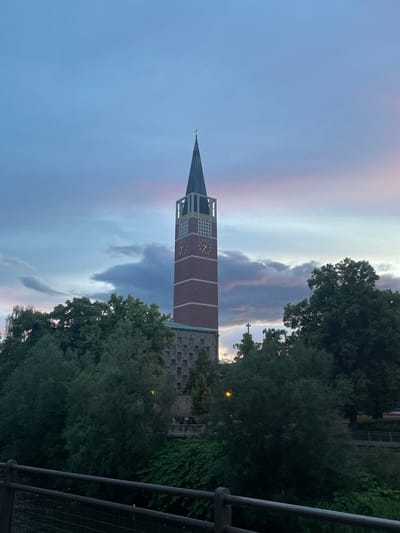 This image captures a tall church tower with a prominent spire against a twilight sky in Pforzheim, Germany. The sky displays soft gradients of blue, pink, and purple, interspersed with dark, dramatic cloud formations.

The church tower is constructed of reddish-brown brick, featuring lighter-colored sections with square cut-outs and golden clock faces, each displaying Roman numerals, though the specific time cannot be clearly read. A slender cross caps the very top of the spire. To the right of the tower, an adjacent stone building is partially visible, characterized by numerous small, square, recessed windows and another cross on its roofline.

Dense green trees frame the lower and right portions of the scene, some obscuring a faintly glowing street lamp, which confirms the evening or dusk setting. A dark metal railing, likely part of a bridge or elevated viewpoint, is visible along the far right side of the frame. No people or discernible activity are present, conveying a tranquil and atmospheric scene. The weather appears partly cloudy.
