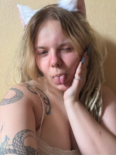 A young Caucasian woman with blonde hair, dreadlocks, and white cat ears is shown in a close-up, head-and-shoulders shot. She is winking and sticking out her tongue, with her left hand resting on her cheek. She is wearing a light-colored lace bra. Tattoos are visible on her left arm and shoulder, including a snake design. She is also wearing a ring with a light-colored stone on her right ring finger. The background is a textured yellow wall. The scene appears to be indoors, possibly in a home.