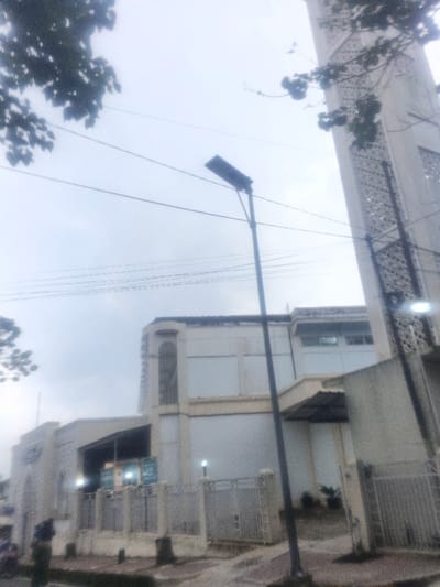 An exterior photograph captures a building complex in Morrumbala, Mozambique, under an overcast sky. The dominant structure on the right is a tall, slender minaret, white with an intricate geometric lattice pattern, extending vertically from the mid-right to the top-right frame. Adjacent to the minaret, a multi-story building with a light-colored facade and a flat roofline features arched design elements and multiple windows, some tinted. A black, rectangular solar-powered streetlight stands centrally, extending from the lower middle to the upper middle, with multiple parallel electrical wires strung behind it. The foreground presents a paved surface, likely a sidewalk, bordered by a light-colored metal fence with concrete posts, featuring two gate sections, one partially open on the left. A partially obscured figure in dark attire is visible behind the left fence section, near a parked vehicle, possibly a scooter. Several illegible signs are mounted on the building facade behind the fence. Dark foliage from trees is visible in the upper left and upper right corners of the frame.