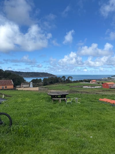 The image displays an elevated view of the Muxía, Spain coastline under a partly cloudy blue sky. The foreground consists of vibrant green grass, featuring a partial black bicycle wheel on the far left. Slightly right of center, a rustic, round wooden picnic table with an attached bench is present. To its right are two dilapidated metal chairs, one painted green. On the far right, three orange pipes are stacked horizontally on the grass. In the midground, a cluster of buildings is visible, including an orange-roofed house on the left and a small light-colored building with a white, multi-faceted roof structure behind it. A dark paved road borders the orange house. Beyond these structures, a dense line of green trees and shrubs separates the land from the ocean. The background reveals the blue Atlantic Ocean, characterized by whitecaps, and a forested landmass forming a bay on the left. Distant red-roofed buildings are visible along the far shoreline to the right. No people or overt actions are depicted.