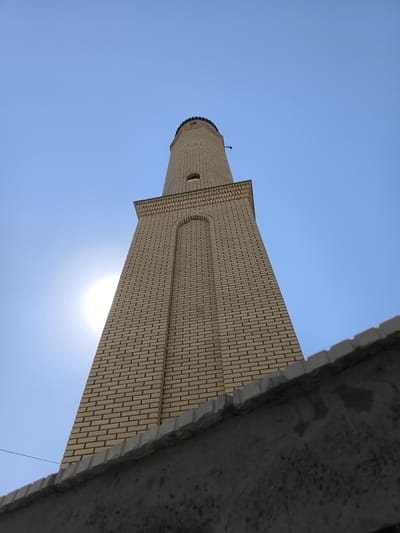 An upward-angle shot shows a tall, narrow minaret made of light-colored bricks against a clear blue sky. A portion of the sun is visible behind the minaret, casting a glare. The minaret has several levels, with distinct architectural details such as arched windows and decorative brickwork near the top. At the very top, a dark, rounded cap is visible. In the foreground, the top of a rough, gray concrete wall runs horizontally across the bottom of the frame, partially obscuring the base of the minaret and creating a sense of depth. A thin black wire is visible stretching across the scene from the left side.