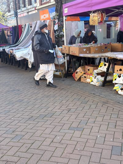 An outdoor market scene in Rotherham, United Kingdom, captured during daylight hours. The foreground features a light brown and grey paved pedestrian walkway laid in a herringbone pattern.
In the mid-ground, an elderly male, appearing of South Asian descent, with a prominent white beard and a grey knit beanie, black sunglasses, a dark grey and black winter jacket, white shalwar-style trousers, and black shoes, walks from left to right. He holds a white plastic bag in his left hand and a dark walking stick in his right hand.
Behind him, to the left, is a market stall displaying various textiles and fabrics draped over a metal frame. Further right, under a purple canopy, is another market stall. Two females, also appearing of South Asian descent, stand behind the stall, examining goods. The female on the left wears a light grey headscarf and a patterned dark top, while the female on the right wears a dark hooded jacket. The stall table is laden with numerous open cardboard boxes, many appearing to be repurposed banana cartons, filled with small items such as trinkets or craft supplies. Empty and collapsed cardboard boxes are visible on the ground beneath this stall.
Visible in the background are traditional brick buildings, typical of a UK town centre, including a facade with an orange and blue sign displaying "WOW VAPE." A tree trunk with artificial flower decorations is present on the far right. A sign on the purple market canopy reads "Facebook wowvape." The scene is illuminated by natural light, indicating a clear day.