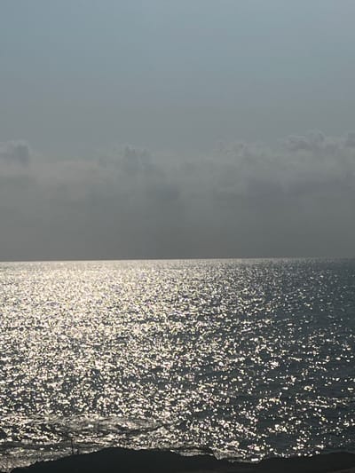 A wide-angle, eye-level shot captures a vast expanse of shimmering ocean under a hazy, overcast sky. The water's surface is a dynamic mosaic of light reflections, creating a sparkling effect as sunlight pierces through the clouds. The horizon line is sharp, dividing the textured water from the muted tones of the sky. In the foreground, a dark, rough edge of land, possibly a coastline or breakwater, juts into the frame from the bottom, its texture contrasting with the smooth water. The location is identified as the city of Cabo Cervera-Playa La Mata, Spain, though no specific landmarks or inhabitants are visible in this serene, naturalistic scene.