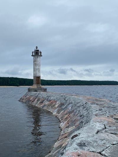 This is a photograph of a lighthouse situated on a stone pier extending into a body of water. The lighthouse is cylindrical and made of concrete, with a weathered appearance and a small entrance door at its base. It is topped with a lantern room. The pier is constructed from large, irregular stones of varying colors, including pinkish-red and gray. The water is dark and somewhat choppy, reflecting the cloudy sky. In the background, a dense green forest lines the opposite shore, with a strip of sand visible along the waterline. The sky is overcast with gray clouds. The scene is located near the settlement of Моторное, Russia. There are no people visible in the image.