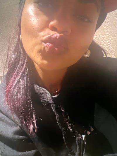 A close-up, upward-angle selfie shows a person with dark skin and dark hair with purple highlights pursing their lips, as if for a kiss. They are wearing a black hoodie and a black headscarf. A sparkly earring is visible on their right ear. The background is a textured, light-colored wall. The lighting suggests the photograph was taken indoors with a light source from the front, creating highlights on the subject's face. The image appears to be a casual self-portrait. There is no indication of the city of Boone, United States of America, within the image itself.