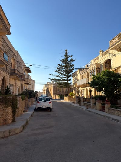 The image depicts a street scene in Xagħra, Malta, under a clear blue sky. On the left, a stone wall borders a building with arched windows and balconies. A blue sign with white lettering reading "KAVARNA" is affixed to this building. A white car is parked on the right side of the street, facing away from the viewer, with its license plate partially visible. Across the street, another building features ornate balconies and decorative elements. A tall, slender pine tree stands prominently in the middle ground, with its branches reaching towards the sky. The street itself is paved with asphalt, and a concrete sidewalk runs alongside the buildings. The overall impression is of a quiet, sun-drenched street in a Mediterranean town.