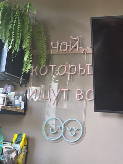 In the city of Brilliant, Russia, an indoor scene depicts a wall adorned with a neon sign and a hanging fern. The neon sign, crafted from pink tubing, is mounted on a clear acrylic board and features text in Cyrillic script. The text reads "Чай, который ищут все," which translates to "Tea that everyone is looking for." Below the text, two intertwined circular outlines of blue neon form smiley faces, representing a whimsical touch.
To the left of the sign, a shelf is visible, holding various items including jars, bottles, and bags of what appears to be coffee or tea. A portion of a microwave is also visible above the shelf. A large fern hangs from the top left of the frame, its fronds cascading downwards. To the right of the neon sign, a black computer monitor is partially visible, its dark screen contrasting with the illuminated text. The overall environment suggests a cafe or a domestic setting with a focus on beverages.