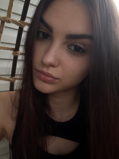 This is a close-up, headshot photograph of a young woman. She has long, dark brown hair that falls around her face and shoulders. Her skin is fair, with some visible pores and a few small blemishes on her cheek. She is wearing a black top with a cutout detail on the chest. A thin silver necklace is visible around her neck. Her eyes are dark and her eyebrows are well-defined. She has a slight pout on her lips.
The background appears to be a textured surface, possibly a wall or a piece of furniture, with vertical lines. To the left of the woman, there is a rope detail, suggesting the presence of a swing or a hammock. The lighting suggests an indoor or shaded outdoor setting.
There is no information available to place this image in Polena, Bulgaria.