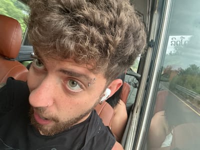 A man with light hair, light eyes, and fair skin takes a selfie in what appears to be a vehicle. He has a nose ring and eyebrow piercings. The man is wearing a black shirt. He's holding the camera at an unusual angle, making the image slightly disorienting. He has a short beard and mustache. The background shows part of the vehicle's interior with brown seats. A window is also visible.