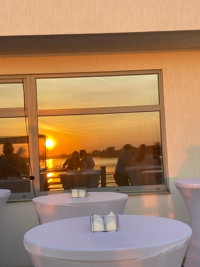 A sunlit room shows a window reflecting an outdoor sunset scene. People are reflected sitting at outdoor tables. There are also white-covered tables with small lanterns in the room. The location may be Aramil, Russia.