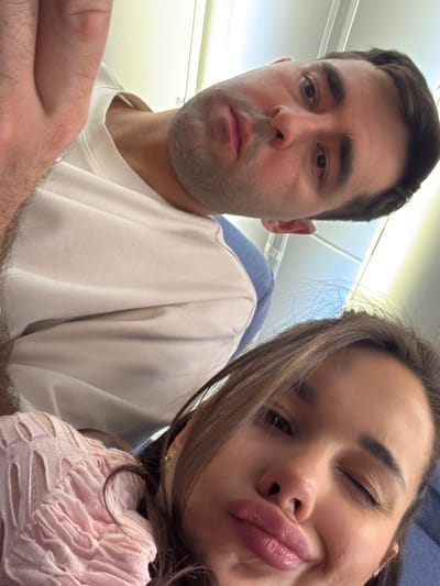A selfie photograph shows two people, presumably a couple, seated on an airplane. The man on the left has dark hair and appears to be wearing a white t-shirt. His expression is somewhat neutral. The woman on the right has long brown hair and is wearing a pink, textured sweater. She is making a kissing face toward the camera. The setting is a passenger aircraft cabin, as indicated by the overhead lights and the blue airline seat visible behind the man. The location in Shunyi District, China, is not discernible from the image.