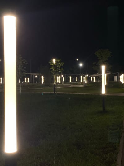 This is a nighttime outdoor photograph of a park in the city of Pokhvistnevo, Russia. The scene is illuminated by numerous tall, vertical, pillar-shaped lamps that emit a bright yellow light. These lamps are distributed throughout the park, lining pathways and areas of grass. In the foreground, a prominent lamp is partially visible on the left side of the frame. In the middle ground, more lamps are arranged in rows, some in pairs. In the background, trees are visible, some with leaves illuminated by the lamps, and a building or shelter structure with a white roof is also present. The ground is covered in grass. The overall atmosphere is calm and softly lit by the artificial lights.