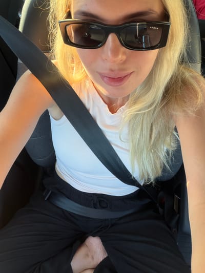A woman with blonde hair and sunglasses is seated in the driver's seat of a vehicle, wearing a white tank top and black pants. She is wearing a seatbelt. Her left arm is resting on the armrest. The image appears to have been taken in the city of Requena, Spain, given the sunlight and the style of the car interior.
