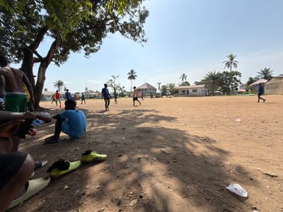 This full-shot image captures a vibrant outdoor scene on a sunny day in Monrovia, Liberia, centered around a casual soccer (football) game. The setting is a wide, unpaved dirt field, appearing dry and dusty, suggesting a period without recent rain. In the background, several modest buildings with various roof types, including corrugated iron, are visible, indicating a community or residential area. Lush green foliage and several tall palm trees dot the landscape, contributing to the tropical ambiance.

The main activity is a group of young men and boys playing soccer. They are scattered across the mid-ground of the field, some actively engaged in play while others walk or stand. Their attire consists of t-shirts and shorts, with some wearing sports jerseys, including one participant on the left side of the field clearly sporting a blue jersey with "3 CHELSEA" visible on the back. An orange cone, possibly marking a goal or drill point, is on the field.

In the immediate foreground, several individuals are resting or observing the game. To the bottom left, a person is seated, holding a smartphone, with a water bottle nearby. Another person sits on the ground with their back to the viewer, wearing a blue t-shirt and shorts. A pair of bright yellow-green soccer cleats lie on the dusty ground in the foreground, alongside various sandals and slides. Large shadows from a prominent, leafy tree on the left stretch across the foreground and part of the playing field, indicating a high sun, likely midday or early afternoon. The sky is bright and clear, with few clouds. The overall mood appears relaxed and communal, centered on the recreational activity. Pieces of debris, such as a white plastic bag, are scattered on the ground.