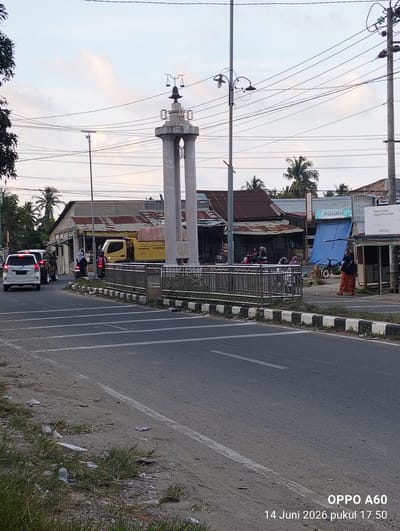 A street scene in Uleepulo, Indonesia, features a landmark monument topped with a scale, flanked by two pillars. Several vehicles, including a white SUV and a yellow truck, are present on the asphalt road, which is marked with white lines. Pedestrians are seen near the monument and the businesses lining the street. The sky above is partly cloudy. Buildings with corrugated metal roofs are visible along the roadside, some displaying signage such as "NVIDIA GROSIR". A blue tarp is draped over a structure. The ground near the road is littered with debris and grass. The image is timestamped "14 Juni 2026 pukul 17.50" and displays "OPPO A60" in the lower right corner.