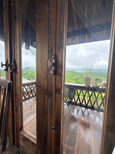 The image shows a view from inside a wooden structure, looking out onto a wet wooden balcony and a lush green, hilly landscape under a cloudy sky. The interior features wooden walls and door frames, with visible copper pipes running vertically along the walls. One of these copper pipes has a decorative switch mechanism attached. A wooden chair is partially visible on the left. The balcony has a railing with a criss-cross pattern. On the balcony, a small potted plant sits on a saucer, accompanied by decorative figurines of animals. The overall scene suggests a tranquil, potentially rural setting in Thôn 2, Vietnam, likely a balcony or porch area of a dwelling or guesthouse.