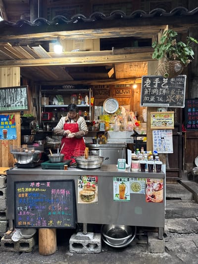 This image depicts a street food stall operating within a traditional wooden structure in Gongtan, China. A single individual, appearing to be an adult with short dark hair and wearing a red apron over a light-colored top, stands behind a stainless steel counter. They are actively engaged in preparing food, holding and shaping a small, light-colored dough-like item with both hands.

The stall's interior wall is constructed from dark wood planks and features intricately carved wooden panels. Overhead, exposed wooden beams support a roof with dark, traditional tiles, and a single bare light bulb hangs, illuminating the workspace. A framed picture depicting a black-and-white forest scene hangs on the left wall, above a blue poster advertising "Sour Plum Soup." A woven basket containing a green plant hangs from an upper beam on the right.

The stainless steel counter is the primary workspace, supported by concrete blocks and a wooden stump. On the left side of the counter, a cooking burner with a pot is visible, alongside a metal grate holding several spherical items over heat. Towards the center-right, a deep-fryer pan contains a utensil, and various bottles, jars, and plastic containers are arranged. Two green QR code stickers for payment are affixed to the counter.

Multiple menus and promotional posters are displayed. A large black chalkboard menu leans against the front of the counter, listing items like "Cool Summer," "Osmanthus Fermented Rice Milk Tea," and "Fresh Meat Pancake" with prices. Above the counter, another black sign reads "Old Craftsmanship," and lists "Fresh Meat Pancake" and "Ice Jelly." Additional colorful laminated posters on the counter's front feature images of burgers/sandwiches, fruit drinks, and desserts.

The ground in front of the stall is dark and uneven. To the right, stone steps ascend alongside the structure. Below the counter, several stainless steel bowls are stored.