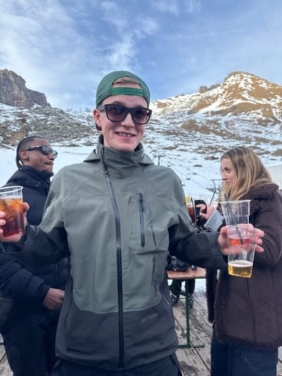 The image captures three individuals enjoying drinks outdoors on a bright, snowy day in a mountainous region, likely Alagna Valsesia, Italy.
In the foreground, a young man with light hair, wearing a green baseball cap backward and dark sunglasses, smiles broadly at the camera. He is dressed in an olive green and dark grey technical jacket, bearing the "ARC'TERYX" logo on the left chest area. He holds two clear plastic cups; one contains an iced, reddish-orange drink with a straw, and the other holds an amber-colored beverage.
Behind him, a woman with long, light brown hair is visible, wearing a brown, possibly fur-lined, winter jacket. She appears to be looking down or away from the camera, and her hand holds a smartphone. Partially visible in the lower right of the frame is a third person, a man in dark sunglasses and a black jacket, holding a clear plastic cup with a dark, iced drink.
The background reveals rugged, snow-dusted mountains under a clear blue sky with sparse white clouds. Structures of a ski lift are discernible between the subjects and the distant peaks, indicating a ski resort environment. The individuals are positioned on what appears to be a wooden deck or terrace. The overall mood is relaxed and social, suggesting an après-ski gathering or a break during a day on the slopes, with the bright sunlight and winter attire pointing to a crisp, cold day.