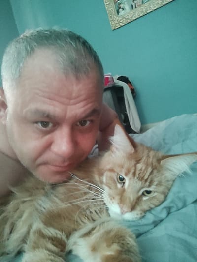 A man and an orange cat are lying on a bed. The man is in the foreground, his head close to the camera, with his left arm visible and resting near the cat. The cat is nestled against his chest, its head slightly turned to the side and its eyes are open and looking towards the camera. The man has short, graying hair and appears to be looking into the camera with a relaxed expression. The cat is long-haired with distinctive markings. The background shows a teal-colored wall with a framed picture hanging on it, and a blurry stack of clothing to the right of the man. The overall lighting suggests it could be daytime, possibly indoors in a bedroom. The image evokes a sense of comfort and companionship between the man and his pet.