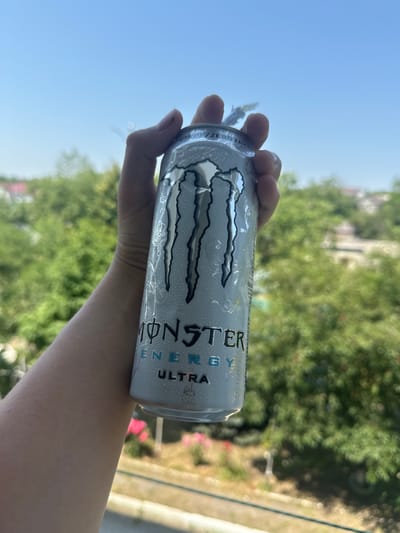 A hand holds a silver Monster Energy Ultra can, with the iconic claw mark logo prominently displayed. The can appears slightly damp, suggesting it has been chilled. The background is a blurred vista of lush green trees and buildings, under a bright blue sky. The presence of greenery and typical residential architecture suggests an outdoor setting, possibly a balcony or window overlooking a neighborhood.