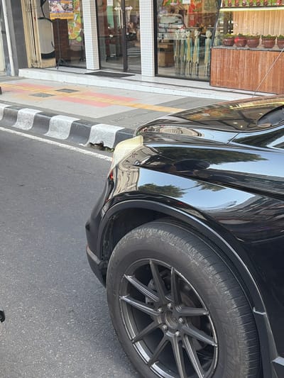 A black SUV is parked on a street in front of a commercial establishment. The vehicle's front fender, wheel, and a portion of its hood are visible. The street is paved with asphalt, and there is a curb with black and white stripes separating it from a sidewalk. The sidewalk features patterned tiles in yellow and pink. Behind the sidewalk, a building with large glass windows and a white-tiled facade is visible. Inside the establishment, mannequins and merchandise can be seen. The scene suggests an urban setting in Magelang, Indonesia, likely during daylight hours.