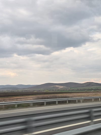 The image presents a panoramic landscape view taken from a moving vehicle on a multi-lane asphalt highway near Yazdach, Bulgaria. The immediate foreground shows the highway's grey surface with white lane markings and two parallel grey metal guardrails, exhibiting motion blur consistent with vehicular movement. Beyond the highway, agricultural fields display sections of dark brown tilled earth and patches of emerging green vegetation. A distant line of light green trees and a faint cluster of structures, indicative of a rural settlement, are visible further back. The background is composed of undulating, vegetated hills, appearing dark, silhouetted against a predominantly overcast sky that features a brighter section near the horizon. No human subjects or explicit actions are depicted.