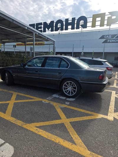 This is a vertically oriented, outdoor photograph taken from a low angle, looking up at a dark gray sedan parked in a lot marked with yellow hazard stripes. The car is positioned on the right side of the frame, with its rear towards the bottom and its front extending towards the top. The car appears to be a BMW, possibly an older model, with silver alloy wheels. The car is parked within a section of the parking lot marked with yellow diagonal lines, indicating a no-parking zone.
To the left of the car, a large building with a white facade is visible. A prominent sign on the building features large, dark letters, the beginning of which reads "ГЕМОХА" (GEMOKHA). Further up on the building, smaller black text on a gray background reads "САД" (SAD) and "РЕМОН" (REMON). Adjacent to the building and partially obscuring it is a metal fence.
The sky is visible above the building and car, with scattered white clouds against a pale blue background. The lighting suggests it is daytime, likely mid-morning or afternoon, with sunlight casting shadows. The overall scene appears to be a commercial or retail area, possibly a large store or shopping center in Sholokhovo, Russia, given the Russian text. No people are visible in the image. The ground is asphalt, with weathered yellow parking lot markings.