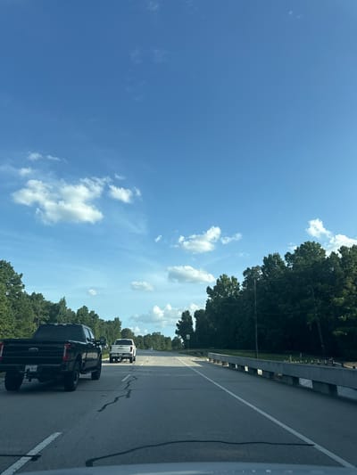 The image captures a view from a vehicle driving on a two-lane road in Magnolia, United States. The road is paved with asphalt and marked with white lines. On the right side of the road, there is a concrete barrier, possibly a bridge railing, with trees and vegetation beyond it. On the left side, there is a black Ford pickup truck in the foreground, followed by a white GMC pickup truck. Both trucks are driving in the same direction. Further down the road, more trees line the landscape. The sky above is a clear blue with scattered white clouds. There are no people visible in the image. The overall scene depicts a typical daytime drive on a suburban or rural road.