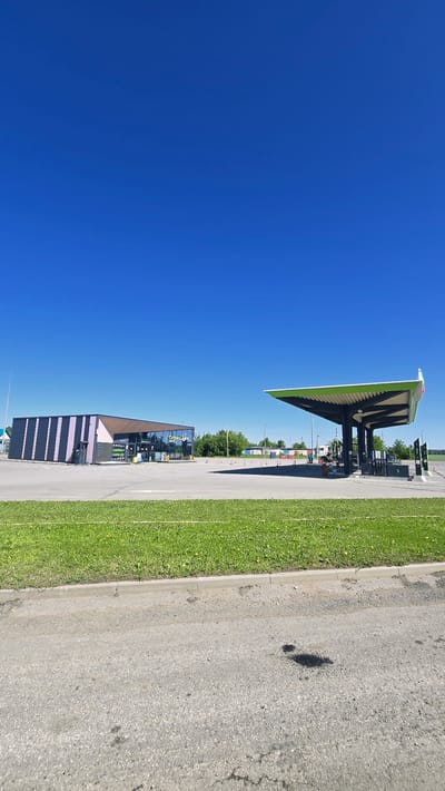 The image captures a bright, sunny day at a gas station in Актюбинский, Russia. The sky is a clear, vibrant blue, dominating the upper portion of the frame. In the mid-ground, a modern gas station complex is visible. To the left, a rectangular building with a dark facade featuring vertical pink stripes and a brown roof houses a convenience store or cafe, with large glass windows. The signage above the windows reads "Green Light Cafe". Adjacent to this building, on the left side, are gas pumps. To the right of the building is a covered gas station canopy with a green roof supported by dark poles. Several gas pumps are situated beneath this canopy. A few vehicles are present at the pumps, and two individuals, one wearing a blue shirt and the other a yellow shirt, are seen near the pumps. The gas station is surrounded by a paved area. In the foreground, a strip of green grass runs along a concrete curb, and beyond that, a grey asphalt road with some dark oil stains is visible. The background consists of trees and some distant structures under the clear blue sky.