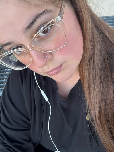 The image is a close-up, selfie-style shot of a young woman wearing clear, cat-eye glasses and a black zip-up hoodie. She has a nose piercing with a small, clear stud. Her long, brown hair is visible, falling over her right shoulder. White earbuds are in her ears, with the cord visible. The background appears to be an outdoor setting, possibly a park or seating area with a metal grate visible behind her left shoulder. The lighting suggests it is daytime. The woman is looking down and to her left, with a slight smile.