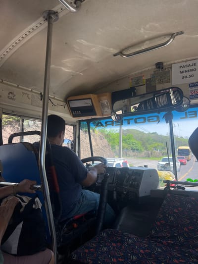 The image shows the interior of a bus, looking towards the front. A male bus driver is seated behind the steering wheel, wearing a dark t-shirt and blue jeans. The bus is in motion, as indicated by the view of traffic and a hillside outside the windows. There are signs on the ceiling of the bus, including one that reads "PASAJE MINIMO $0.50". A camera is mounted above the driver's area. The seats in the bus have a patterned fabric. Outside, there are other vehicles, including a yellow bus and a red car, on a road that appears to be in a rural or suburban area with hills and vegetation. The lighting suggests it is daytime. The image is taken from a passenger's perspective within the bus.