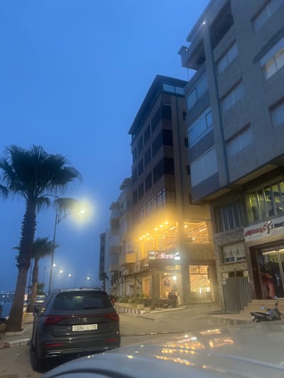 The image depicts a street scene in Fnideq, Morocco. A grey Seat Tarraco SUV is parked on the side of the road, its rear facing the camera. Its license plate reads "13684 75". Palm trees line the street on the left. The street is illuminated by streetlights. Buildings, some brightly lit, flank the street. A sign reading "Café California" is visible above a storefront. The sky is a pale blue. A car is partially visible on the right side of the frame.