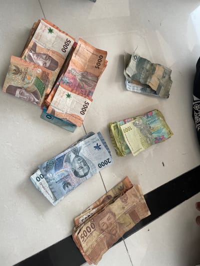 The image displays a scattering of Indonesian Rupiah banknotes on a tiled floor. The denominations visible include 5000, 2000, and 1000. The banknotes are arranged in several piles and spread out across the frame. The floor appears to be light-colored tile with some dirt or debris present. The lighting suggests an indoor environment. There are no people clearly visible in the image, although a hand is partially visible in the bottom right corner. The scene does not provide any specific information that would allow for identification of the location within Trieng Paloh, Indonesia.