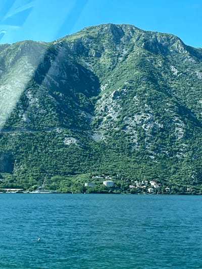 The image shows a mountain covered in dense green vegetation, transitioning to a shoreline with scattered buildings, and finally the blue water of Kotor Bay. The location is likely Kostanjica, Montenegro, given the mountain terrain along the bay. No people are visible. A few boats are docked along the shoreline.