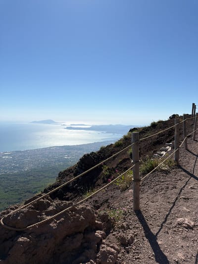 This is a high-angle, medium shot of a walking path on the slope of Mount Vesuvius, overlooking the Bay of Naples and the city of Ercolano, Italy. The path is made of dark volcanic gravel and is bordered by a rope fence with wooden posts. The rope is frayed and worn in places. Small, pink wildflowers grow sporadically along the path. In the distance, the blue water of the bay is visible, dotted with ships, and the coastline of Ercolano stretches along the shore, appearing as a dense urban area with buildings. Further out, islands rise from the sea under a clear, bright blue sky. The scene is devoid of people. The lighting suggests a sunny day, casting shadows from the fence posts onto the path.