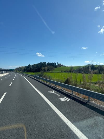 The image displays a multi-lane asphalt highway on a clear, sunny day, located in the vicinity of Ancona, Italy. The foreground features the dark grey road surface with a solid white line on the right shoulder and a dashed white lane separator to its left. White chevron markings, containing the text "100m", are painted on the asphalt to the right of the solid line, indicating a recommended safe distance. A two-bar metallic guardrail runs along the right edge of the highway.

Beyond the guardrail, dry, light-colored vegetation is visible, followed by a wire fence. An expansive, vibrant green agricultural field, likely planted with crops, stretches horizontally across the midground. Rolling hills form the background, adorned with a mix of evergreen and bare deciduous trees. Scattered rural structures are visible on these hills; specifically, a cluster of stone buildings, possibly ruins or an old settlement, is discernible within the green field area to the right. Further back, on a higher hill, more isolated farmhouses or buildings are present among the trees. The faint blue line of the Adriatic Sea is visible on the distant horizon to the right.

Overhead, power lines extend horizontally across the middle of the frame. The sky is a bright, clear blue, dotted with scattered white cumulus clouds. A prominent, elongated aircraft contrail with a diffuse end stretches diagonally from the upper left towards the middle right of the sky. On the far left, an elevated section of highway or an overpass structure is partially visible. No individuals or vehicles are explicitly depicted in the immediate scene, suggesting the perspective is from a moving vehicle.