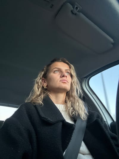A woman is in a car, looking upwards. She wears a black coat, a white sweater, and a seatbelt. Her blonde hair is wavy, and she has a decorative earring. The car's sun visor is visible, as is the windshield and a glimpse of the outside. The photo is taken from a low angle, looking up at the woman.