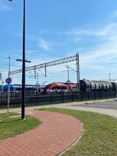 This image depicts a train station with two trains visible. The train on the left is blue and has "MISTRZ MISTRZ" and "KOLEJORZ MISTRZ" written on its side. The train on the right is red and white, with people visible on the platform. Behind the trains, there are large, dark grey cylindrical cargo tanks with "GAJTX" written in white letters. The scene is set against a blue sky with some white clouds. A black lamppost is in the foreground, and a red brick walkway with a bicycle symbol is also visible. The location is the train station in Dobieżyn, Poland.
