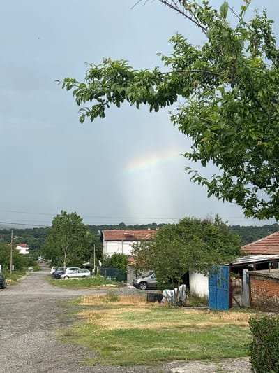 The image shows a rural scene, seemingly in the countryside. A rainbow is visible in the sky, arching behind a tree with green foliage. In the background, there are houses with tiled roofs, and a building with a blue door, indicative of a village setting. Vehicles are parked along a gravel road that runs through the area. A forested area is visible in the distance. The image appears to be taken in or near Cherni Vrah, Bulgaria, based on the provided location.