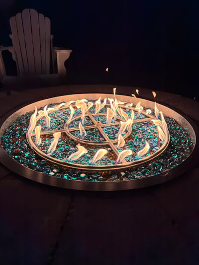 In Flanders, United States of America, a close-up, high-angle shot captures a circular fire pit glowing at night. The fire pit is filled with teal-colored glass rocks and features a star-shaped metal burner from which flames erupt in a dynamic display. The flames are a mix of yellow and blue hues, creating an inviting and warm atmosphere. In the background, a light-colored Adirondack chair is visible, casting a shadow and suggesting a setting for relaxation or outdoor gathering. The surrounding environment is dark, emphasizing the light emanating from the fire pit.