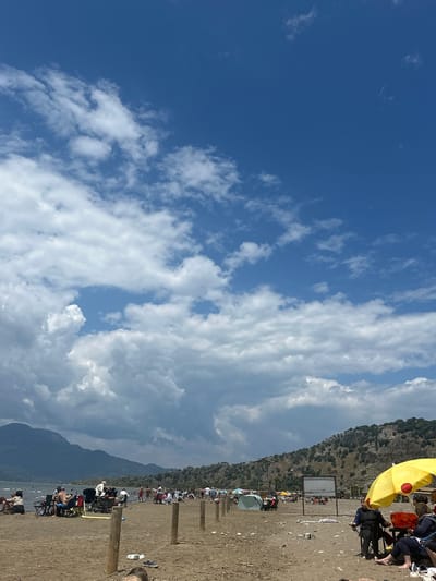 The image depicts a beach scene in Ortaca, Turkey. A mountain range borders the beach. The sky is partly cloudy, with a section of deep blue. The beach is populated with people, many under umbrellas. There is an information board present on the beach. Wooden posts line the beach. People are sitting in chairs along the beach.