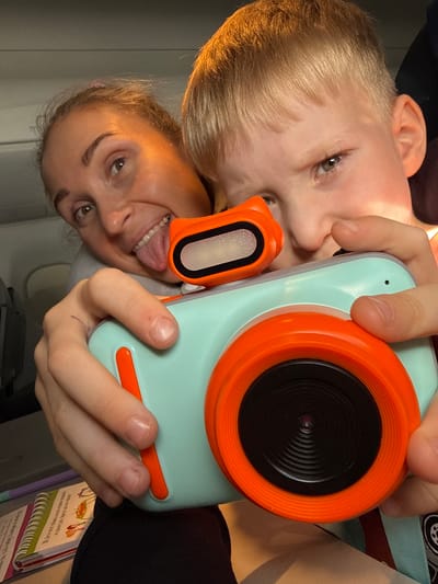 The image shows a close-up selfie of a woman and a young boy, both holding a teal and orange digital camera. The woman is in the foreground on the left, sticking her tongue out and making a playful expression. The boy is on the right, holding the camera with both hands, and looking towards the camera with a slightly mischievous grin. The camera has a large orange lens and a smaller orange component on top, likely a flash or viewfinder. The background is dimly lit and appears to be the interior of an airplane cabin, suggested by the faint lighting and the presence of what might be a seat or a window. There is also a notebook with colorful drawings visible in the lower left corner. The scene captures a moment of shared activity and fun between the woman and the boy. The location of Vykhodnoi, Russia is not visible or inferable from the image.