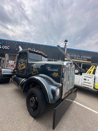 This media file features a black and gold custom Kenworth tow truck parked on asphalt. The truck is adorned with gold pinstriping and decals, including an eagle and the "Smokey and the Bandit" logo. The number "69" is prominently displayed on the front grille area. The truck is positioned in front of a building with a large sign that reads "LUXURY D HOMES FURNITURE" and "VIVE HOME FURNITURE."
In the background, another tow truck, primarily yellow and white, is visible with text indicating "TOWING," "RECOVERY," and "TARE 6360 KG." The sky above is overcast with clouds, suggesting it's a cloudy day. The scene appears to be a commercial area, possibly a business parking lot. The image is taken from a low angle, looking up at the tow truck. The overall impression is one of a well-maintained, customized vehicle at a commercial establishment. The city of Leduc, Canada, is provided as location context.