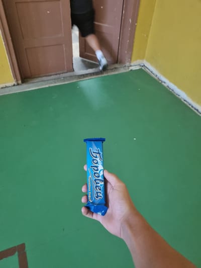 A hand holds a blue wrapper with white text that appears to say "Topoley" and a smaller line of text below it. The wrapper is positioned in the foreground, with the hand holding it from below. The background is a large expanse of green floor and a yellow wall. To the left, a brown wooden door is partially open, revealing a person's legs and feet in dark shorts and white socks, as they are walking away from the camera and through the doorway. The setting appears to be an interior space, possibly a school or gym, given the green floor and yellow walls. The presence of a doorway with a person exiting suggests movement or transition within the space.