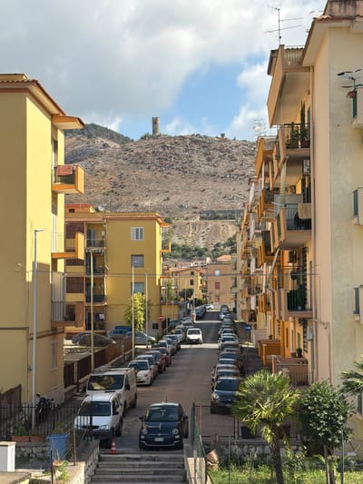 This image captures a residential street scene in Maddaloni, Italy, viewed from an elevated perspective, looking down a street towards a distant, rocky hillside. The original image is rotated 90 degrees clockwise, so the sky appears on the left and the ground on the right.
The scene is dominated by rows of multi-story residential buildings, primarily painted in shades of light yellow and beige. These buildings feature balconies, some with awnings extended, and various windows. Television antennas are visible on several rooftops. The street itself is paved and lined with numerous parked cars and vans on both sides, indicating a densely populated area. Notable vehicles include a dark blue Fiat 500 parked prominently on the right, displaying a partially visible Italian license plate "FW 927NW". Further down the street, a white van has a red traffic cone placed behind it. A blue circular road sign with a white arrow is visible on a pole on the left side of the street (when correctly oriented).
In the background, a large, rugged, and arid hillside or mountain, covered in sparse vegetation, rises dramatically. A small, stone, tower-like structure is visible on a distant peak of this hillside. Green foliage, including palm trees and various bushes, adds natural elements to the urban environment, particularly along the street and adjacent to the buildings.
The time of day appears to be late morning or afternoon, suggested by the bright daylight and clear shadows cast by the buildings and vehicles. The sky is partly cloudy with patches of blue visible. No people are visible, and the overall mood is one of quiet, everyday life in a Mediterranean town setting.