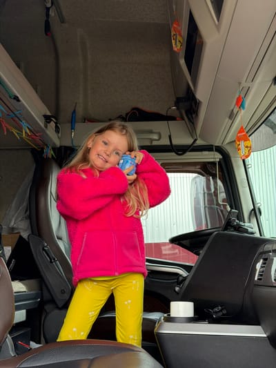 A young girl with blonde hair and blue eyes smiles brightly, looking directly at the camera. She is wearing a vibrant pink fuzzy jacket and yellow pants adorned with white palm tree motifs and the word "ADVENTURE." The girl is lying on her side on a grey and brown seat, holding a blue bottle that has "Macton" and "30" written on it.
The scene is set inside what appears to be the cabin of a large vehicle, possibly a truck or camper, evident from the interior layout, overhead compartments, and a partially visible steering wheel on the right. Daylight filters in through the windows, illuminating the interior. Various personal items and accessories are visible: several air fresheners, including one shaped like a leaf with the brand "AREON" and images of fruit, and two smaller, colorful, mouse-shaped ones. Cables and wires are strung across different surfaces, and a line with colorful plastic clothes pegs can be seen on the left, suggesting a lived-in space. The girl's joyful expression suggests a relaxed and happy atmosphere. The location is Kamyennoye, Russia.