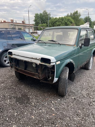 The image depicts a derelict green Lada Niva parked in an outdoor lot in Khar'ino, Russia. The car is heavily damaged, with missing front panels and a detached tire resting against the front fender. The vehicle is showing signs of rust and decay. A dark-colored modern SUV is parked adjacent to the Lada. The surrounding environment is a gravel lot with sparse vegetation and utility poles in the background, suggesting an industrial or semi-urban setting. Buildings are visible in the distance. The sky is overcast, indicating a cloudy day.