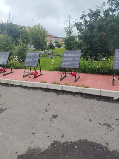 The image shows a memorial in the city of Буланаш, Russia, commemorating individuals. Several black memorial plaques are erected on metal stands, arranged in a line on a red tiled pathway. Each plaque contains a list of names and some associated information, possibly indicating their roles or locations related to their passing. Red flowers are placed at the base of two of the memorial plaques, suggesting recent tributes. In the background, there is a grassy area with trees and shrubs, and behind that, buildings, including what appears to be a residential or public building. The sky is overcast. The foreground shows a wet asphalt surface, indicating recent rain or damp conditions.