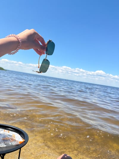 A hand with a pink beaded bracelet and a ring is holding a pair of sunglasses over a body of water. The sunglasses have dark green lenses and gold frames. The water is calm with ripples, and the bottom is visible in the shallows. In the background, there is a clear blue sky with a few white clouds, and a distant shoreline. A small black table with some items on it is visible in the lower left corner. The scene appears to be a waterfront location, possibly a beach or lakefront in Lebedivka, Ukraine.